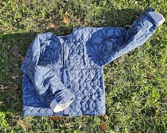 May include: A quilted jacket in shades of blue, featuring a diamond pattern on the body and striped sleeves. The jacket has a white lining at the cuffs and a white zipper closure. The garment is lying on a bed of green grass.