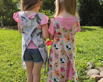 May include: Two children wearing aprons outdoors. One apron is patterned with a newspaper print and dragonfly illustrations. The other apron is light pink with floral seed packet designs. Both children wear pink shirts and denim shorts.