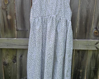 May include: A sleeveless, light-colored dress with a delicate green leaf pattern. The dress is hanging on a white hanger against a weathered wooden fence. The dress has a gathered waist and a flowing skirt.