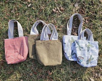 May include: Five small tote bags with different patterns and colors. The bags are made of fabric and have white handles. The patterns include red and white stripes, green corduroy, and a white background with a dragonfly pattern.