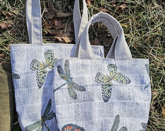 May include: Two tote bags with white fabric and a print of butterflies and dragonflies. The bags have gray canvas straps.