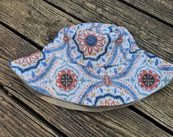 May include: A white bucket hat with a colorful floral pattern in shades of blue, orange, and yellow. The hat has a wide brim and a neutral-colored lining. The hat is made of fabric and is designed for sun protection.