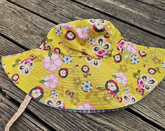 May include: A yellow sun hat with a wide brim, adorned with a floral pattern in pink, blue, and purple. The hat has a quilted texture and a chin strap. The brim measures approximately 12 inches in diameter.