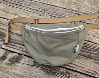 May include: An olive green fanny pack featuring a tan adjustable strap, a white zipper lining, and a metallic D-ring attachment on the front. The bag has a curved, crescent-shaped design with a central zipper closure across the top section.