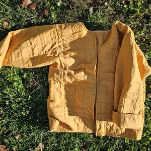 May include: A mustard yellow quilted jacket with a zipper closure. The jacket has a relaxed fit and a slightly oversized look.