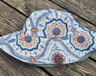 May include: A reversible sun hat with a detailed floral pattern in blue, orange, and white. The hat has a wide brim and a solid green underside. The hat is made of fabric and is designed for sun protection.