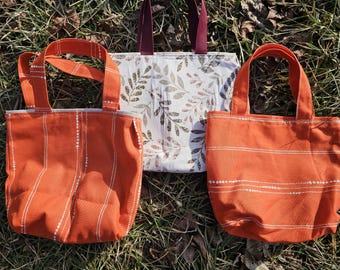 May include: Three tote bags made of fabric. One bag is orange with white stripes, another is orange with white dots, and the third is white with a brown leaf pattern.