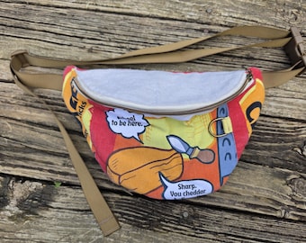May include: A fanny pack featuring a colorful fabric print of cheese, knives, and speech bubbles with the text "to be here." and "Sharp. You cheddar believe it." The bag has a light gray top panel, a zipper closure, and an adjustable tan strap.
