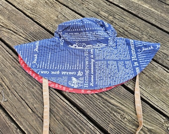 May include: A blue and red sun hat with a wide brim. The hat features a blue fabric printed with text and illustrations, and a red trim. The hat has beige ties and is designed for sun protection.