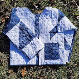 May include: A quilted jacket in shades of white and blue, featuring a geometric pattern. The jacket has a V-neck design, long sleeves, and a patch pocket. The fabric is quilted with a wavy stitch.