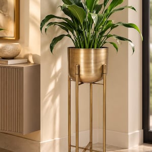 May include: A vibrant green leafy plant potted in a brushed brass-colored metal planter. The planter is supported by a tall, slender, four-legged gold-toned metal stand with a cross-brace design at the base, elevating the plant off the floor.
