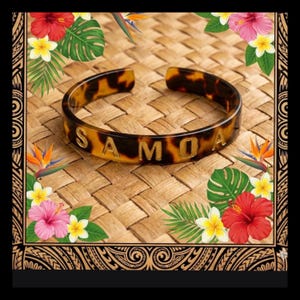 May include: A brown and black patterned bangle bracelet with the word "SAMOA" in gold lettering. The bracelet is set against a woven background and surrounded by tropical flowers and leaves.
