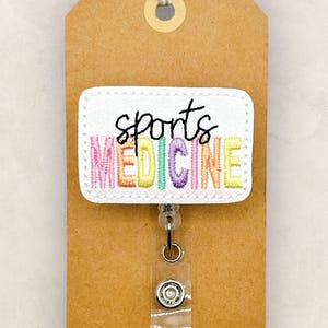 May include: A badge reel with a white rectangular patch embroidered with the words "sports MEDICINE" in colorful letters. The reel is attached to a brown tag with a string.