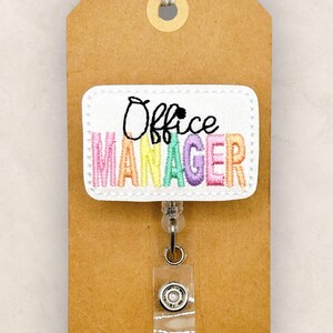 May include: A white badge reel with the words "Office Manager" embroidered in colorful thread. The badge is attached to a brown tag with a string and a clear plastic reel clip.