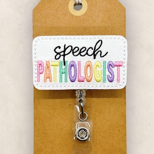 May include: A white badge reel with the words "speech pathologist" embroidered in colorful thread. The badge is attached to a brown tag with a string. The reel has a clear plastic strap and a metal clip.