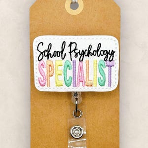 May include: A badge reel with a white embroidered patch that reads "School Psychology Specialist" in colorful letters. The badge is attached to a brown tag with a string at the top. The Small Town Sunshine Company logo is in the upper left corner.