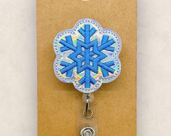 May include: A badge reel featuring a blue embroidered snowflake design on a scalloped, iridescent background. The snowflake is centered on a circular base attached to a clear plastic retractable cord mechanism with a metal snap closure.