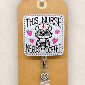 May include: A silver and white badge holder with a gray raccoon wearing a nurse's hat and holding a cup of coffee. The badge holder has pink hearts and the text "This Nurse Needs Coffee".
