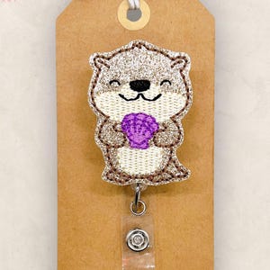 May include: A glittery badge reel featuring an embroidered otter holding a purple seashell. The otter is tan and brown with a black nose and smiling face. The reel is attached to a brown tag with the text "The SmallTown Sunshine Company".