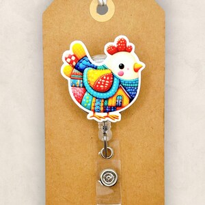 May include: A colorful acrylic badge reel featuring a whimsical chicken design. The chicken is adorned with vibrant patterns in red, blue, yellow, and green. The reel is attached to a brown tag with the text "The SmallTown Sunshine Company" and "Acrylic".
