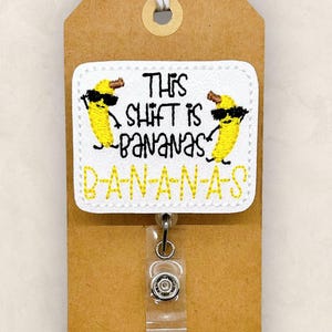 May include: A white rectangular badge reel featuring embroidered yellow bananas wearing black sunglasses. The text reads This shift is bananas B-A-N-A-N-A-S in black and yellow stitching. The badge is attached to a brown card with a white and tan string.