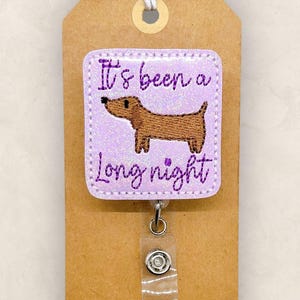 May include: A light purple badge reel with the text "It's been a long night" and a brown dachshund dog illustration. The badge is attached to a brown tag with a string.