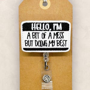 May include: A white and black badge reel with the text "HELLO, I'M A BIT OF A MESS BUT DOING MY BEST" on a brown tag. The reel has a clear plastic clip and a silver-colored metal button. The tag is tied with a natural-colored twine.