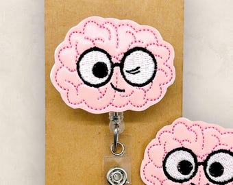 Pode incluir: Um crachá em forma de cérebro rosa e um alfinete a condizer com óculos de armação preta e um olho a piscar. O crachá está preso a uma etiqueta castanha com o texto "The SmallTown Sunshine Company". O alfinete tem uma cara sorridente.