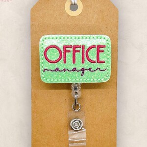 May include: A green and pink Office Manager badge reel. The badge has the words "OFFICE manager" embroidered in pink thread on a green background. The badge is attached to a retractable reel with a clear plastic strap.