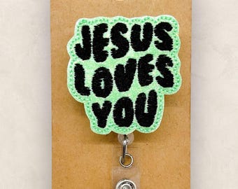 May include: A badge reel featuring a light green, cloud-shaped embroidered patch with the black text 'JESUS LOVES YOU' centered on the front. The patch is attached to a clear plastic retractable cord mechanism mounted on a brown cardstock tag.