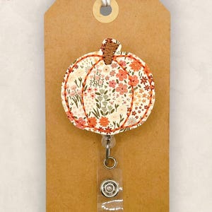May include: A pumpkin-shaped badge reel featuring a cream-colored fabric with a floral pattern in shades of orange, pink, and green. The pumpkin has a brown stem and is outlined with orange stitching, attached to a clear plastic clip.