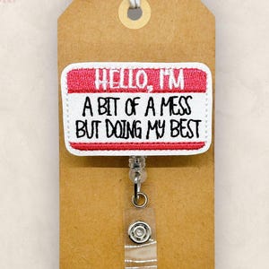 May include: A badge reel with a white and red name tag design that reads "HELLO, I'M A BIT OF A MESS BUT DOING MY BEST." The badge is attached to a brown tag with a string.