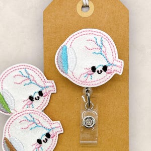 May include: Embroidered eye badge reel and two eye patches. The eye designs feature white, pink, blue, and brown details with a smiling face. The badge reel is attached to a brown tag with a string. The text "small wnau shin designs" is at the top left.