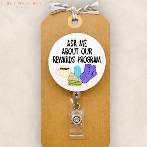 May include: White round badge with a black and white image of a coffee cup, a sandwich, and a pair of purple socks. The text "Ask me about our rewards program" is printed in black on the badge.