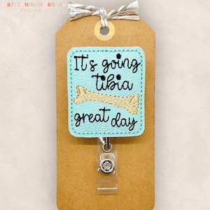 May include: A light blue badge reel with the phrase "It's going tibia great day" embroidered in black, featuring a bone graphic. The badge is attached to a brown tag with a white and tan string.