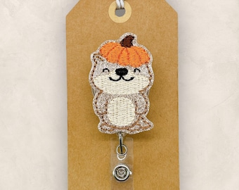 May include: A badge reel featuring an embroidered otter wearing an orange pumpkin on its head. The otter has light brown fur, a cream-colored belly, and a black nose and mouth. It is attached to a clear plastic clip on a brown paper tag.