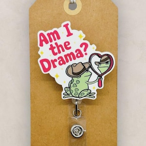 May include: A badge reel with a cartoon frog wearing a cowboy hat and looking in a heart-shaped mirror. The text "Am I the Drama?" is above the frog. The badge is attached to a brown tag with a twine bow.