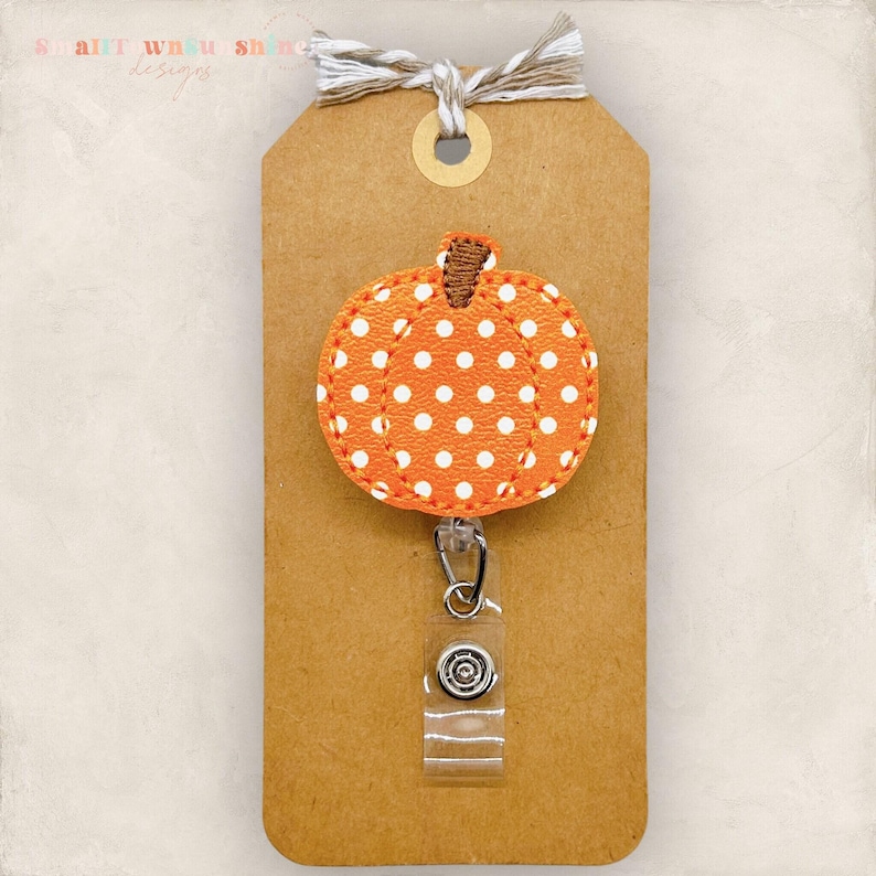 May include: An orange pumpkin-shaped badge reel with white polka dots, attached to a brown tag. The tag has a hole at the top with a twine bow. The reel has a clear plastic clip.