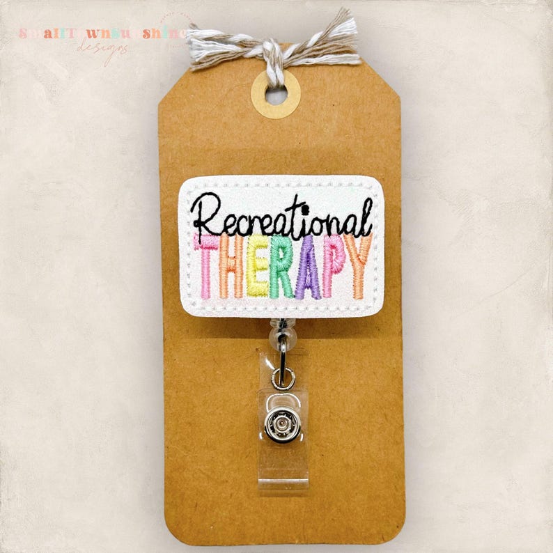 May include: A white badge reel with the words "Recreational THERAPY" embroidered in colorful thread. The badge is attached to a brown tag with a string. The reel has a clear plastic clip.