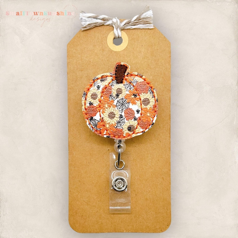May include: A pumpkin-shaped badge reel with a fall floral design. The reel features a brown stem and a pattern of orange, yellow, and white flowers with black spiderwebs. It is attached to a brown tag with a twine bow.