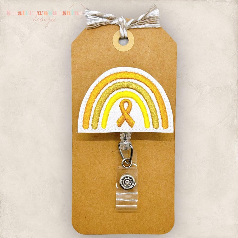 May include: A badge reel with a yellow and white rainbow design and a yellow awareness ribbon. The badge reel is attached to a brown tag with a string. The text "Smalltown Sunshine Designs" is visible in the upper left corner.