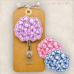 May include: Three hydrangea-shaped badge reels featuring embroidered floral patterns. One light purple badge reel is attached to a brown paper tag with a white string. Two additional loose badge reels in pink and blue are positioned nearby.