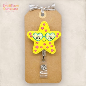 May include: A yellow star-shaped badge reel with a smiling face and heart-shaped glasses. The star has pink polka dots and is attached to a brown tag with the text "The SmallTown Sunshine Company". The tag is tied with twine.