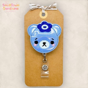 May include: A light blue bear-shaped badge reel with a dark blue flower on its head. The bear has a sad expression with pink cheeks and black eyes. The badge reel is attached to a brown tag with the text "The Small Town Sunshine Company".