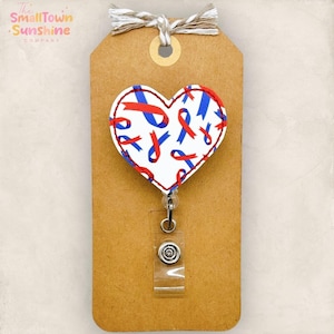 May include: A heart-shaped badge reel with a white background and red and blue ribbon pattern. The reel is attached to a brown tag with the text "The Small Town Sunshine Company". The tag is tied with twine.