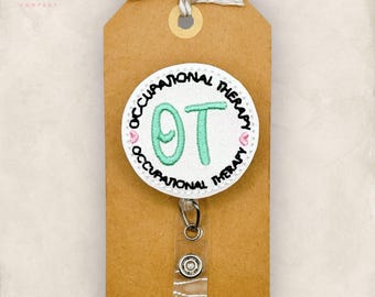 May include: A round badge reel featuring white fabric with black embroidered text reading OCCUPATIONAL THERAPY and large mint green embroidered letters OT. Two small pink hearts are stitched on the sides. The reel is attached to a brown card.