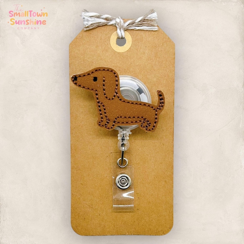 May include: A brown dachshund badge reel with black stitching, attached to a brown tag with a white and tan string. The badge reel has a clear plastic strap and a silver-colored clip.