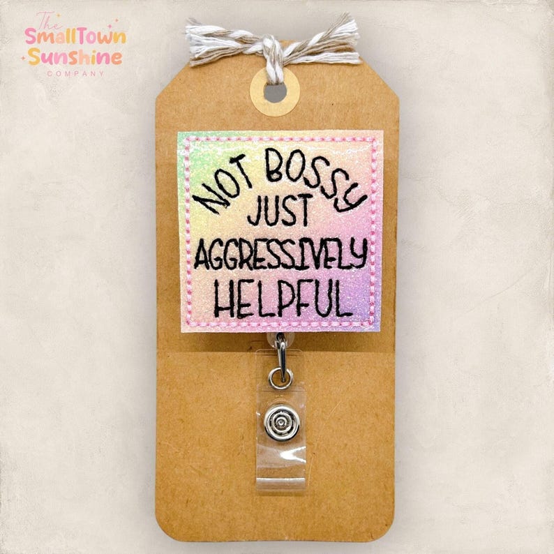 May include: A brown tag with a badge that reads "NOT BOSSY JUST AGGRESSIVELY HELPFUL". The badge has a colorful, glittery background and a pink border. A retractable badge reel is attached.