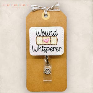 May include: A white badge reel with the words "Wound Whisperer" embroidered in black. The design includes a bandage with a pink heart in the center. The badge is attached to a brown tag with a decorative string.