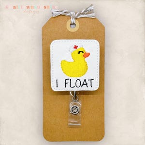 May include: A badge reel with a white square patch featuring a yellow rubber duck wearing a nurse's cap with a red cross. The text "I FLOAT" is below the duck. The reel is attached to a brown tag with a twine bow.